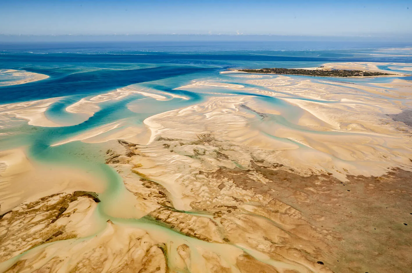 Turquoise tidal channels braiding through wide white sandbanks out towards deep blue open water, seen from the air.